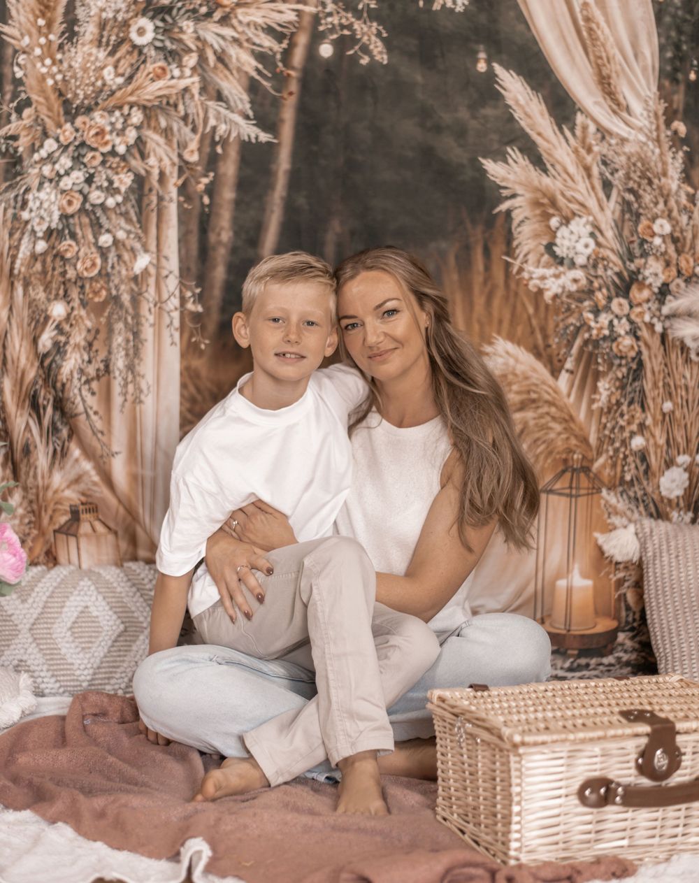 A woman and young boy in white outfits sit together on a pink blanket surrounded by boho floral decor.