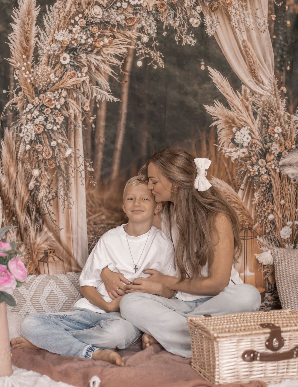 A woman kisses a young boy on the cheek as they sit together in front of a boho floral backdrop.
