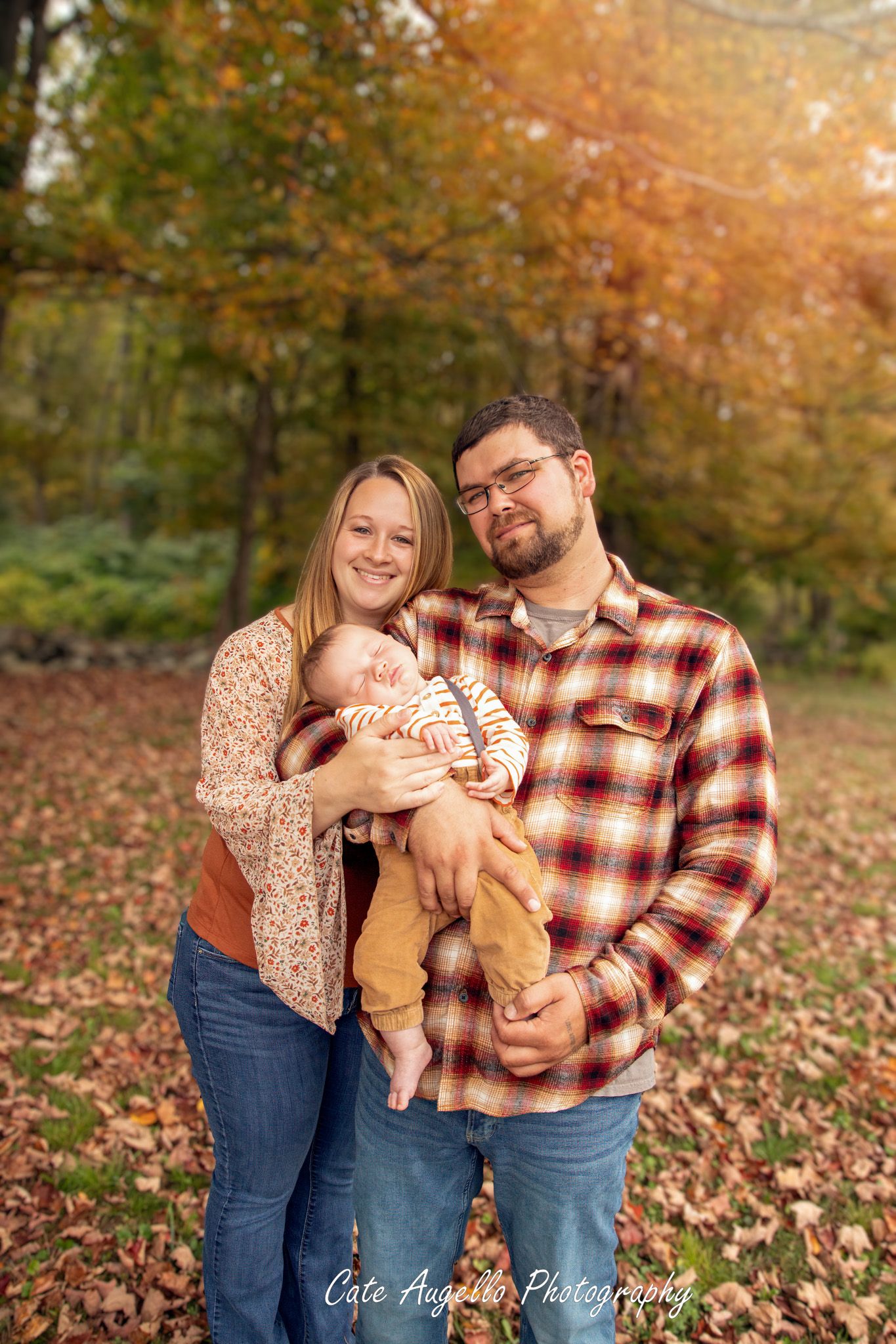 A couple and baby pose together in a scenic autumn forest setting with orange leaves strewn on the ground.  Hebron, CT.  Rainbow baby