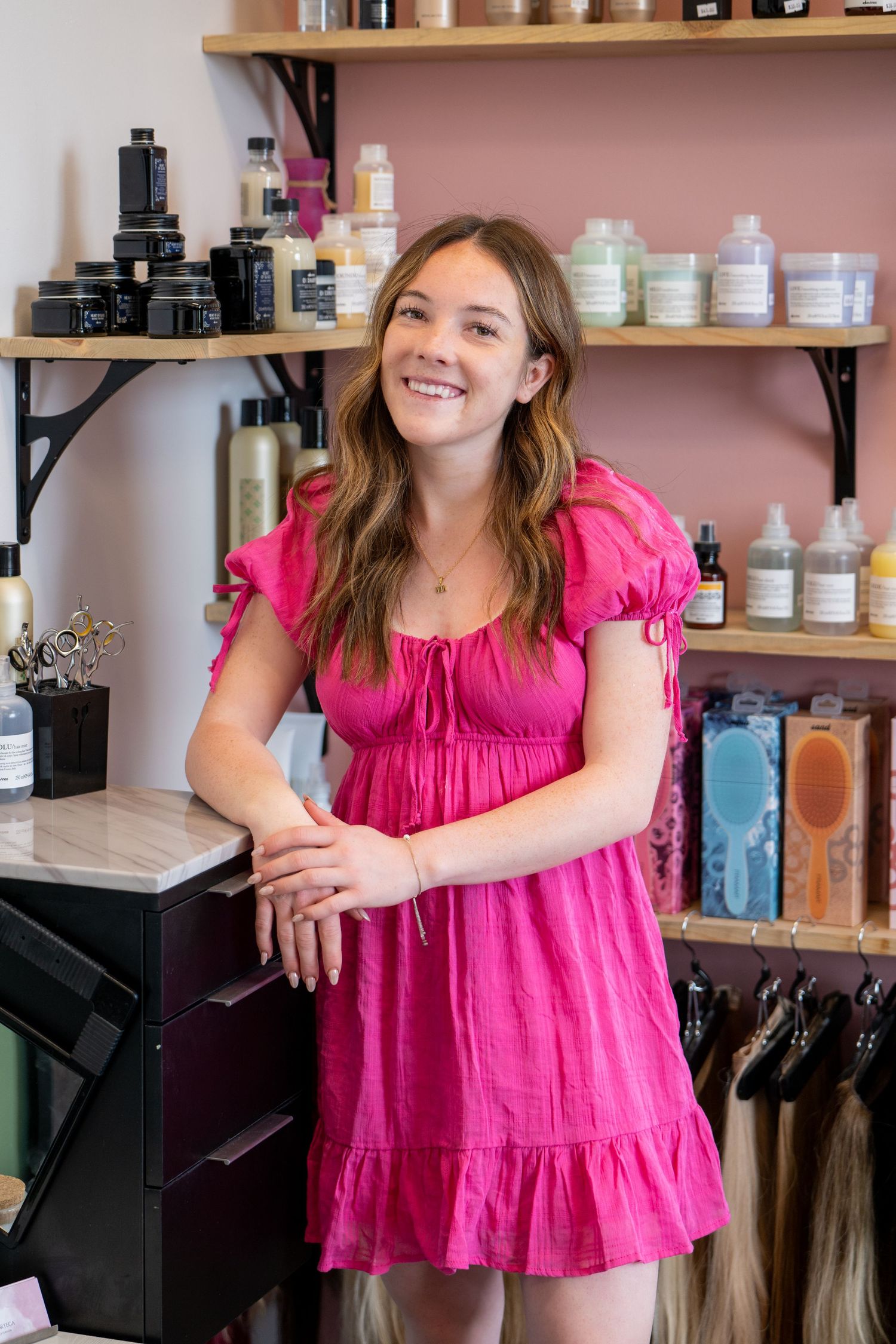 A retail employee in a pink dress stands in front of shelves stocked with beauty products.