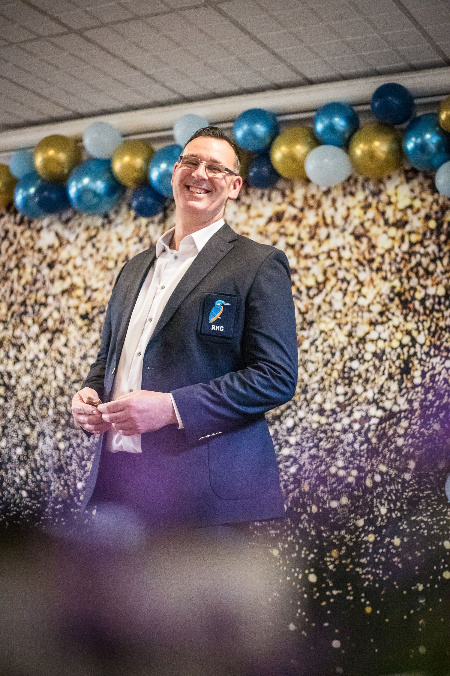 Happy smiling man wearing a Redditch Hockey Club blazer with blurred flowers in the foreground and balloon in the background