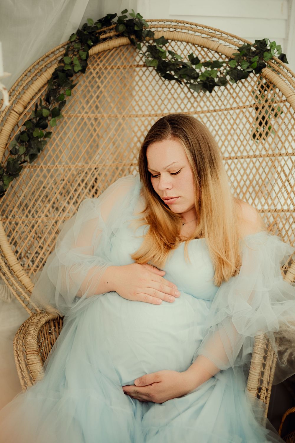 Expecting mother in a wicker chair cradling her pregnant belly and flowy dress
