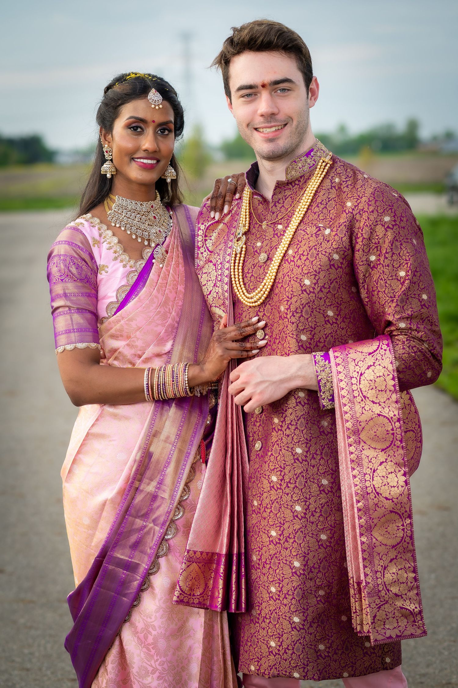A couple in traditional Indian wedding attire poses together outdoors.