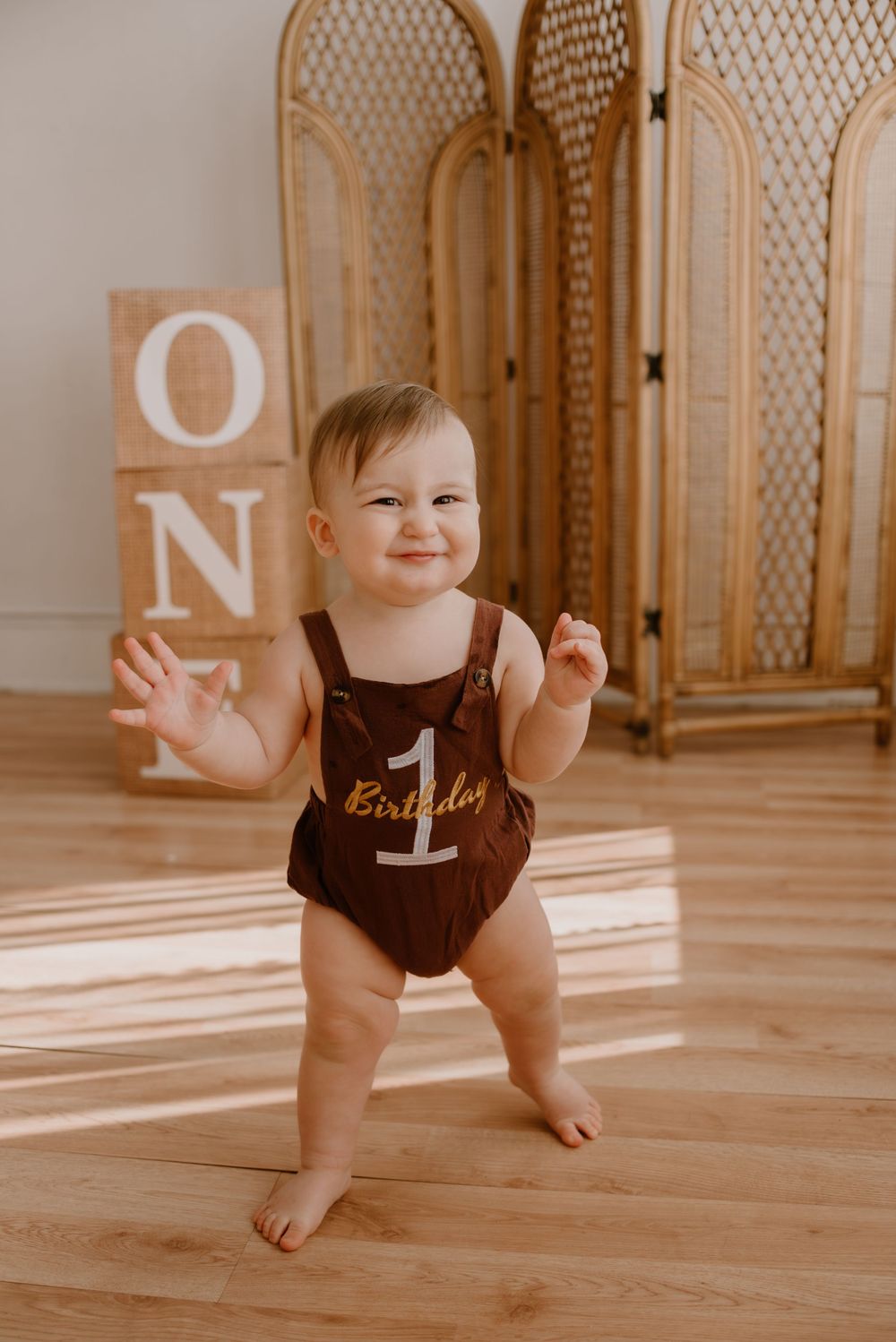 A toddler in a brown romper standing in front of wooden room dividers and decorative blocks spelling 'ONE'.
