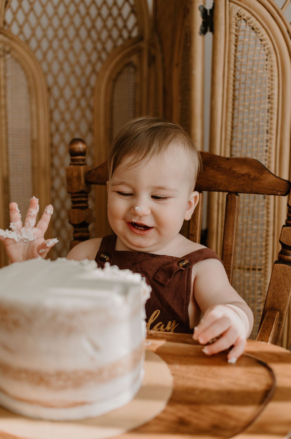 A smiling baby in a brown outfit reaching for a white birthday cake while sitting in a wooden chair.
