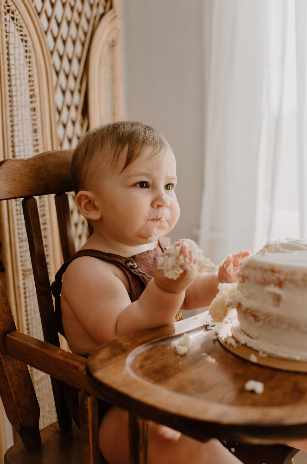 A baby enjoying cake while seated in a wooden chair with a decorative screen in the background.