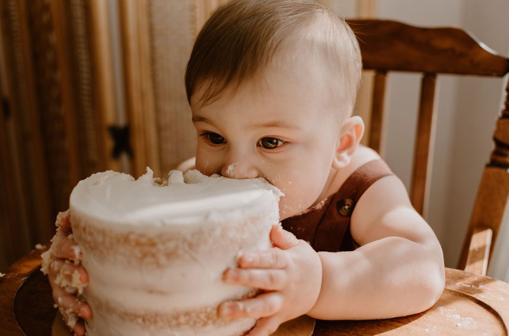 A baby eagerly leaning in close to a white frosted cake while sitting in a wooden high chair.