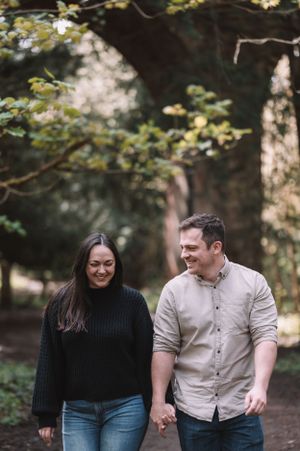 A couple walking hand in hand and laughing together on a wooded trail during autumn.