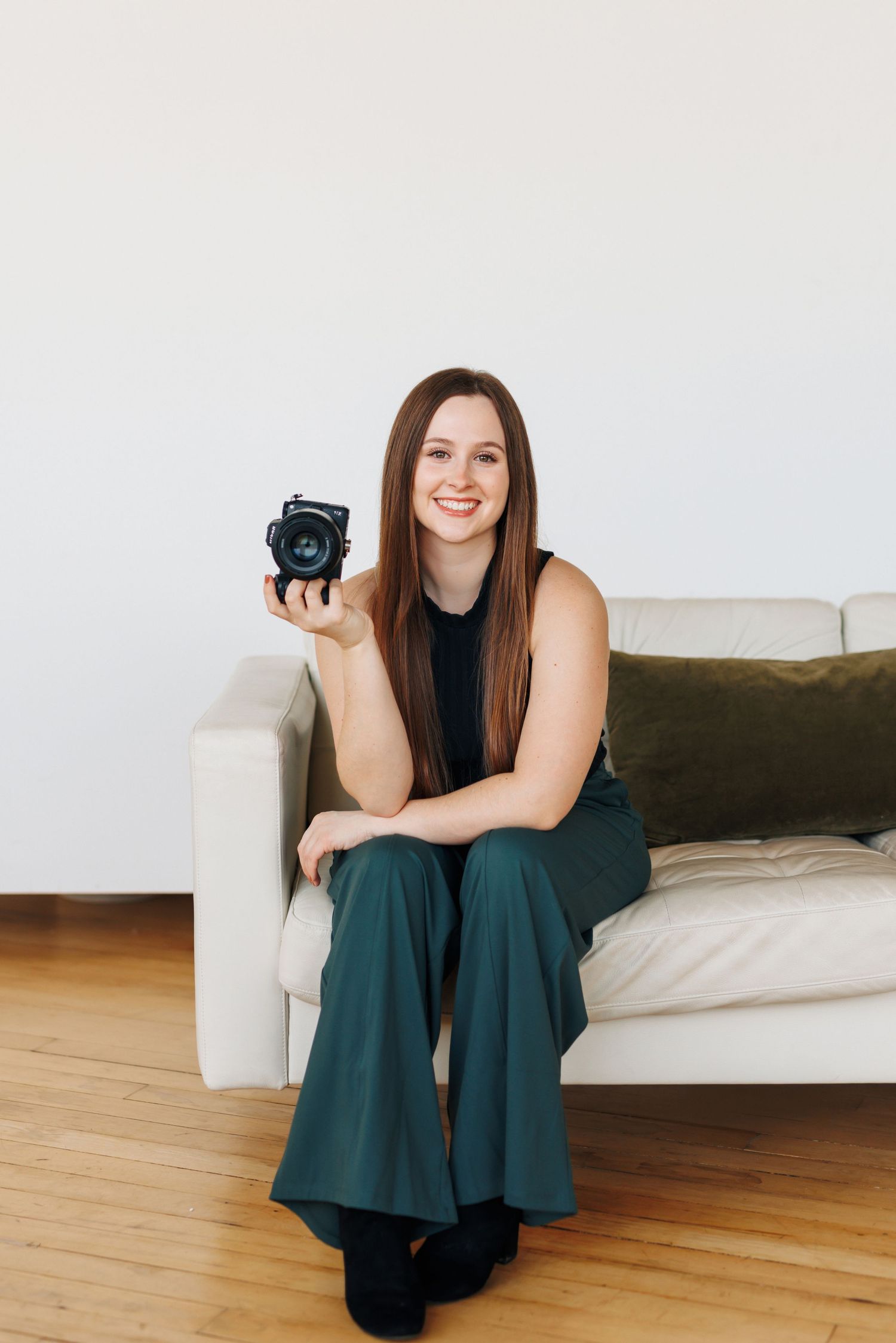 A woman in casual attire sits on a white couch holding a camera.