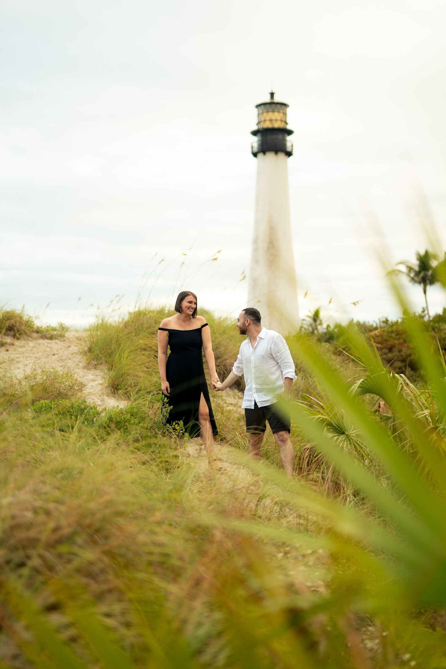 Romantic wedding photos taken near a lighthouse with tall grass in the foreground.