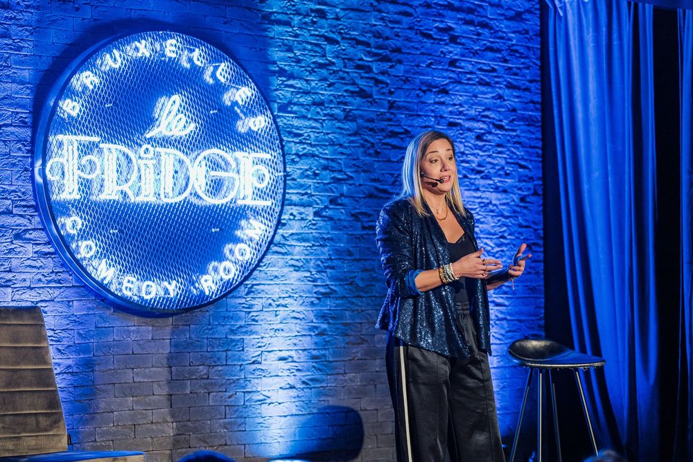 A performer stands on stage at a comedy club with a blue neon 'Fridge' sign glowing on the brick wall behind them.