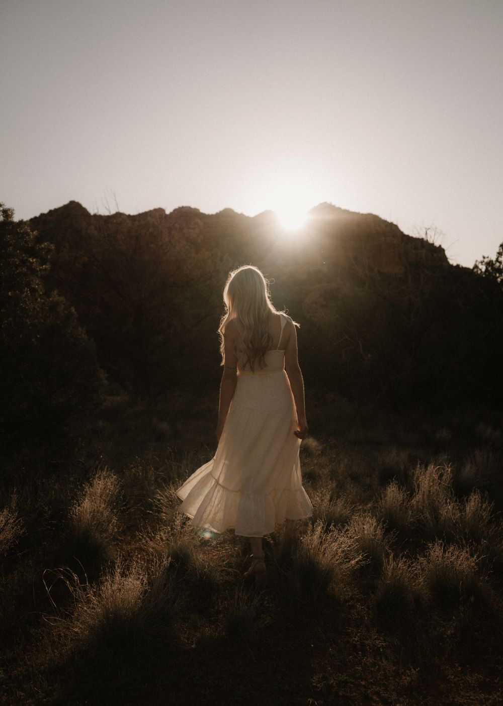 A backlit silhouette in a flowing dress stands against a sunset mountain landscape.