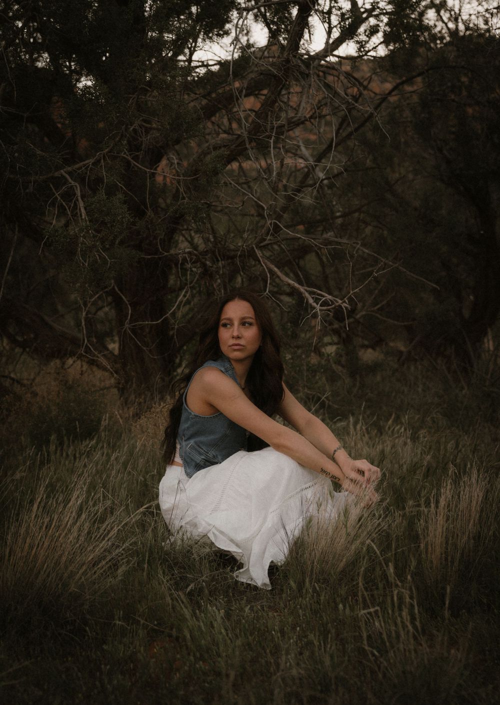 A moody outdoor portrait featuring a flowing white skirt and teal top against a dark natural background.