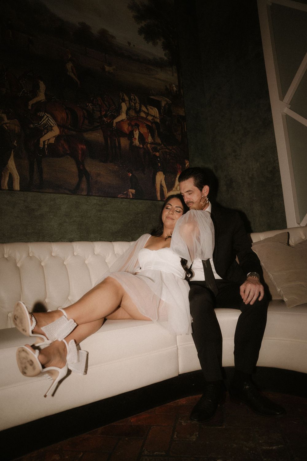 A romantic moment on a white couch beneath vintage artwork in warm lighting.