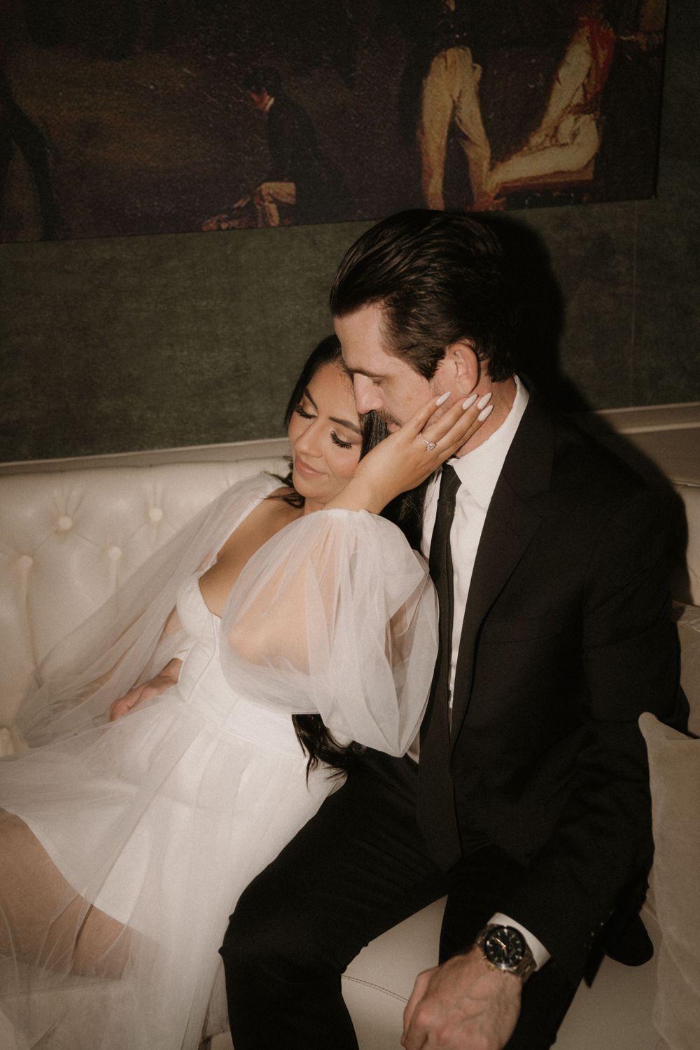 An intimate embrace on a white couch beneath classical artwork in sepia tones.