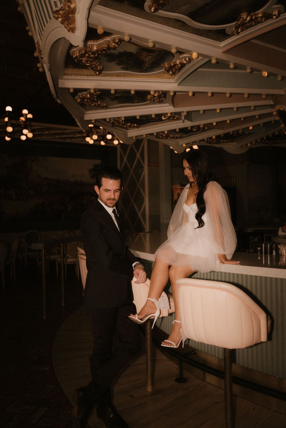A stylish couple poses playfully in a vintage-inspired venue with ornate ceiling details.