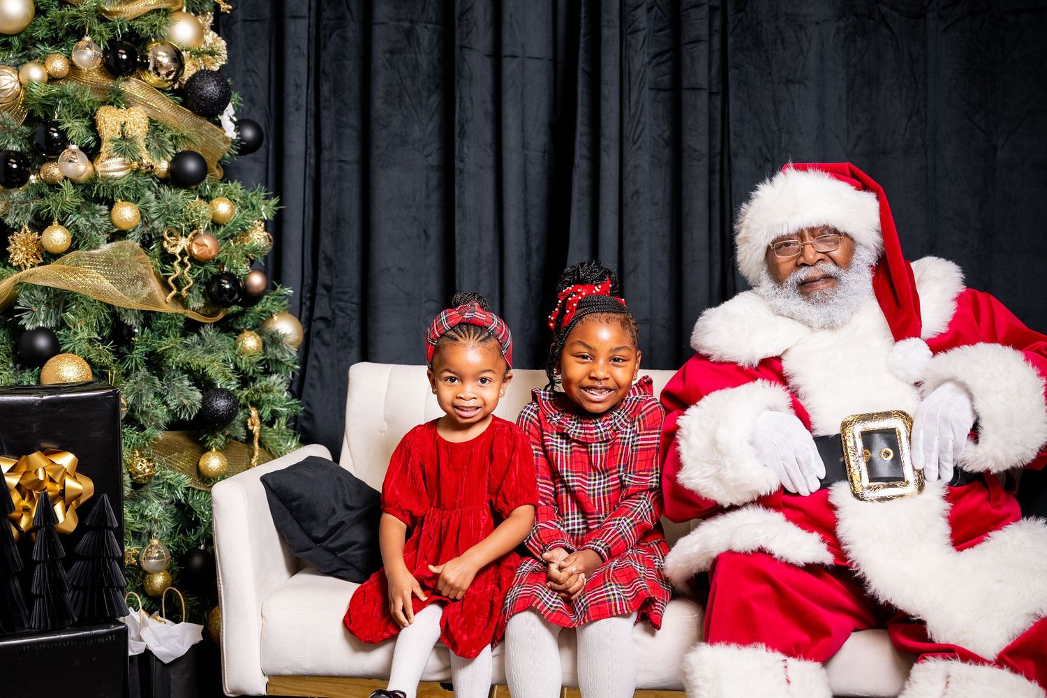 Children in red holiday outfits visit with Santa Claus in front of a decorated Christmas tree during festive photo session.