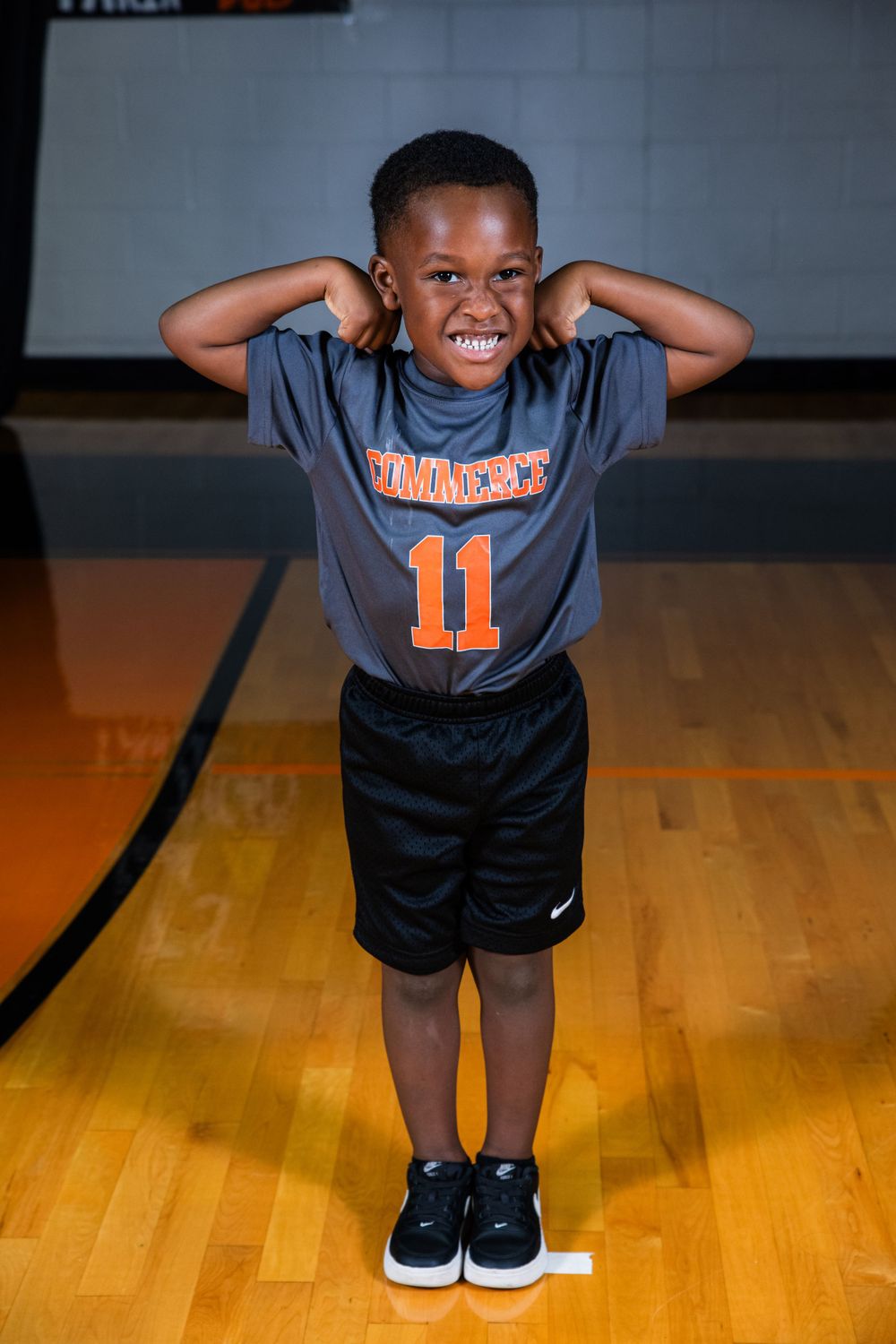 Young athlete in grey #11 jersey and black shorts poses joyfully on a basketball court with wooden flooring.