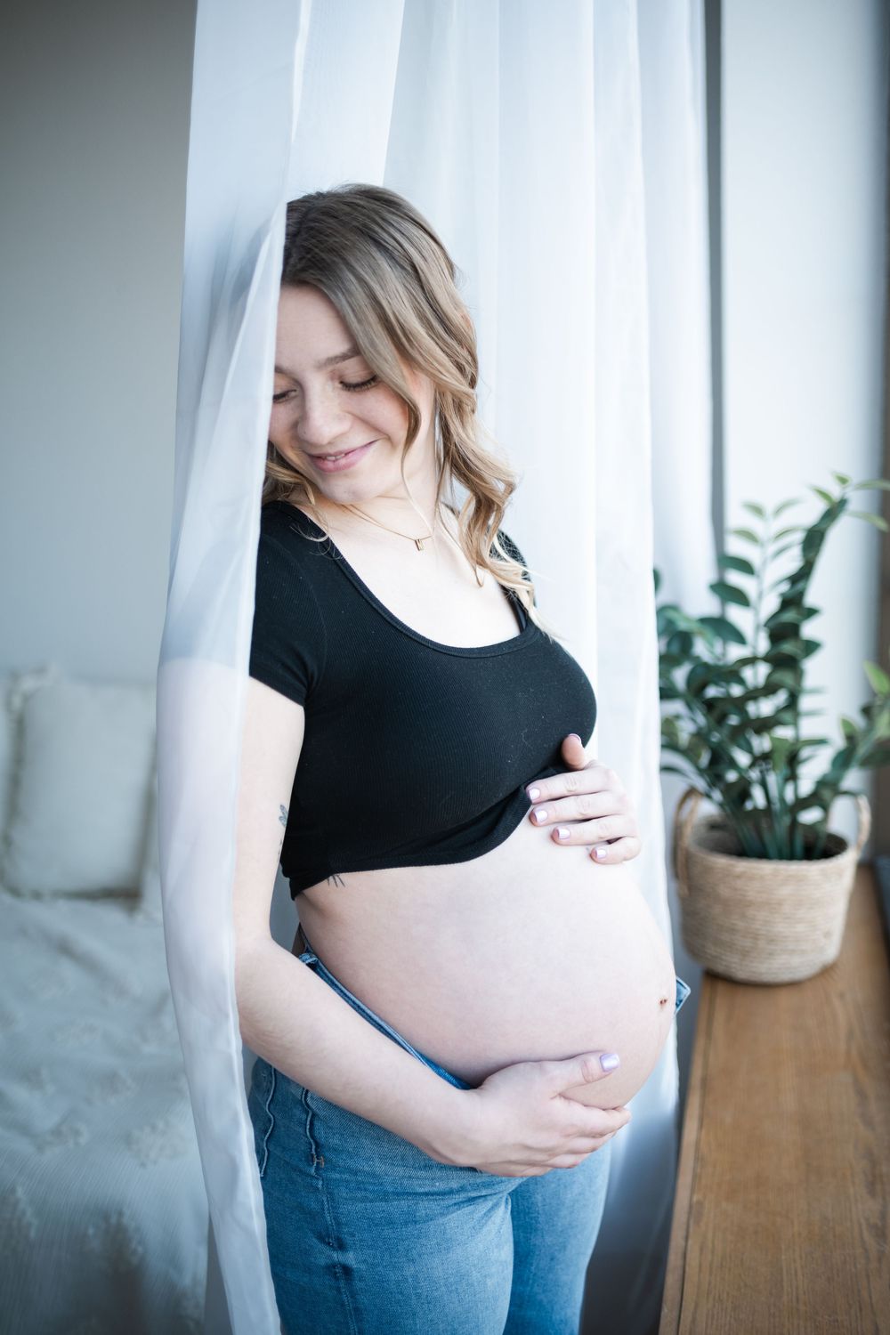 Smiling pregnant woman in black top and jeans cradling her bare baby bump while standing by a white curtain.