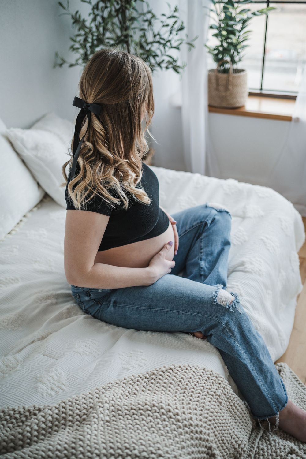 Pregnant woman in black top and ripped jeans sitting on white bed, cradling baby bump, with plants in background.