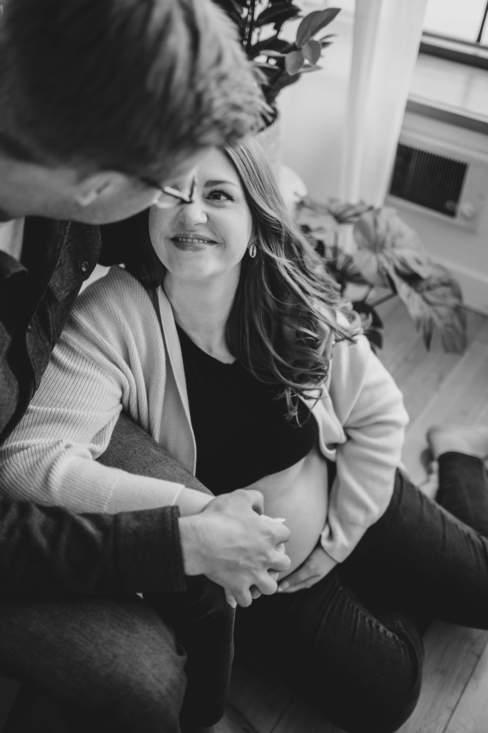 Black and white photo of a smiling pregnant woman sitting with her partner, both holding her baby bump indoors.