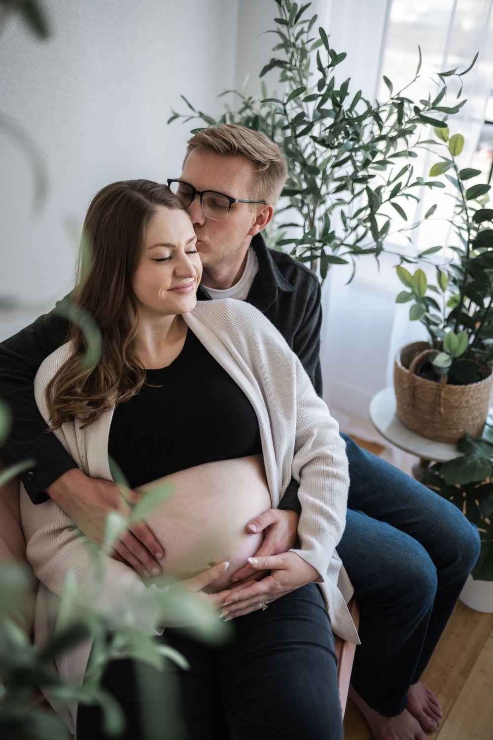A couple tenderly embraces, the man kissing his pregnant partner's head while both cradle her baby bump indoors.
