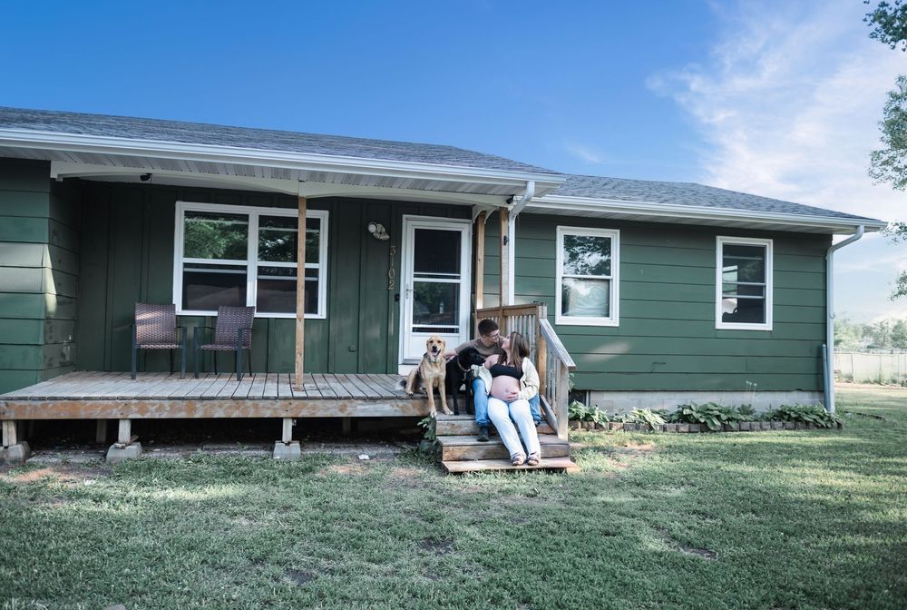 Couple sitting on wooden porch steps of green ranch-style home with dog on sunny day.