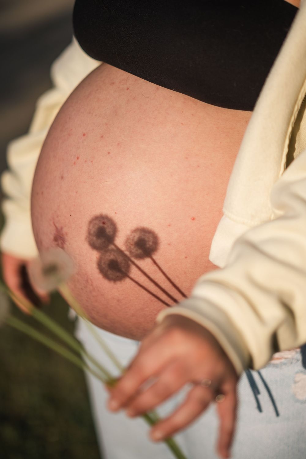 Pregnant woman's bare baby bump with dandelion tattoo, holding wildflowers, wearing jeans and white jacket outdoors.