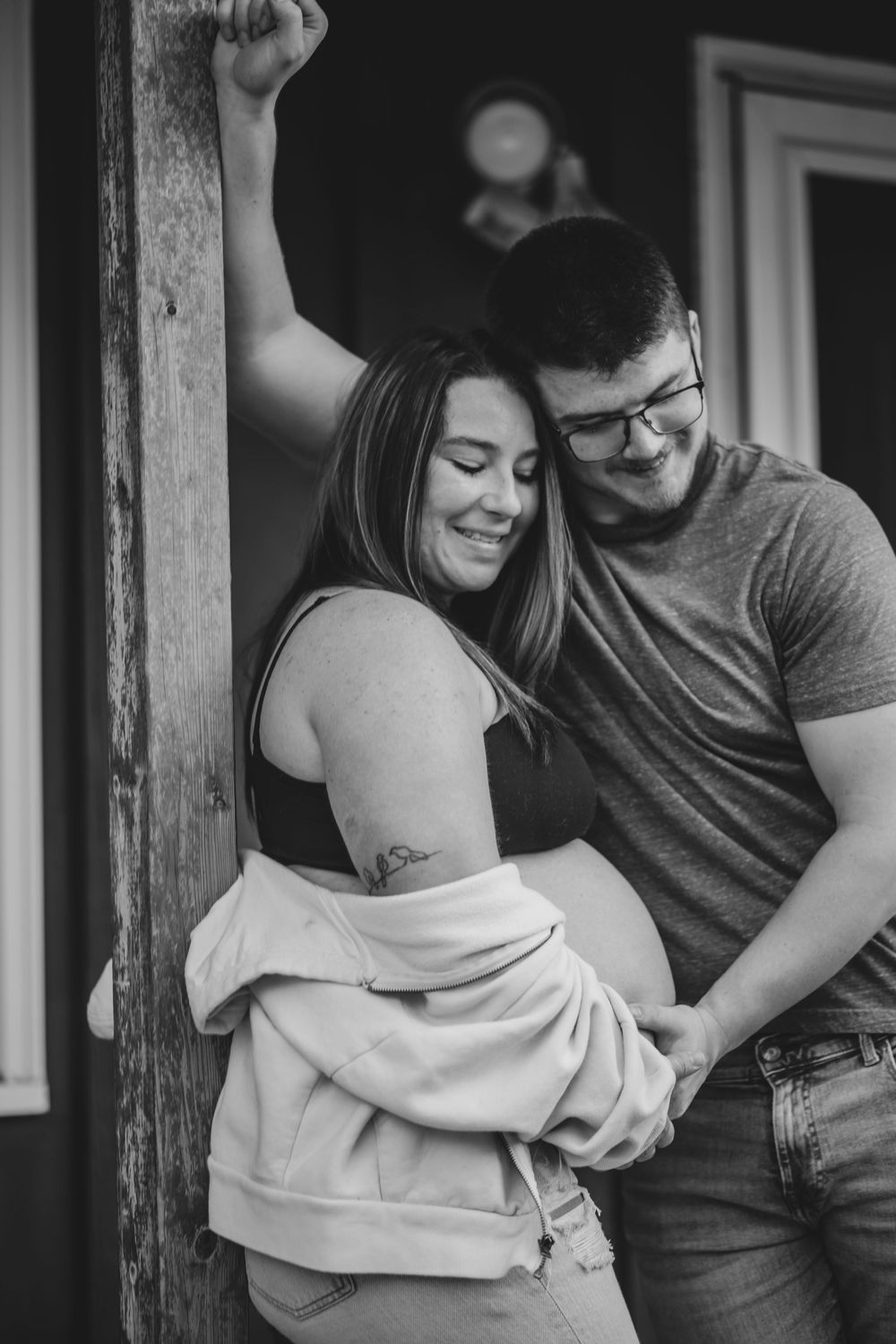 Black and white maternity photo of smiling couple embracing outdoors, man resting chin on womans head while holding her baby bump.