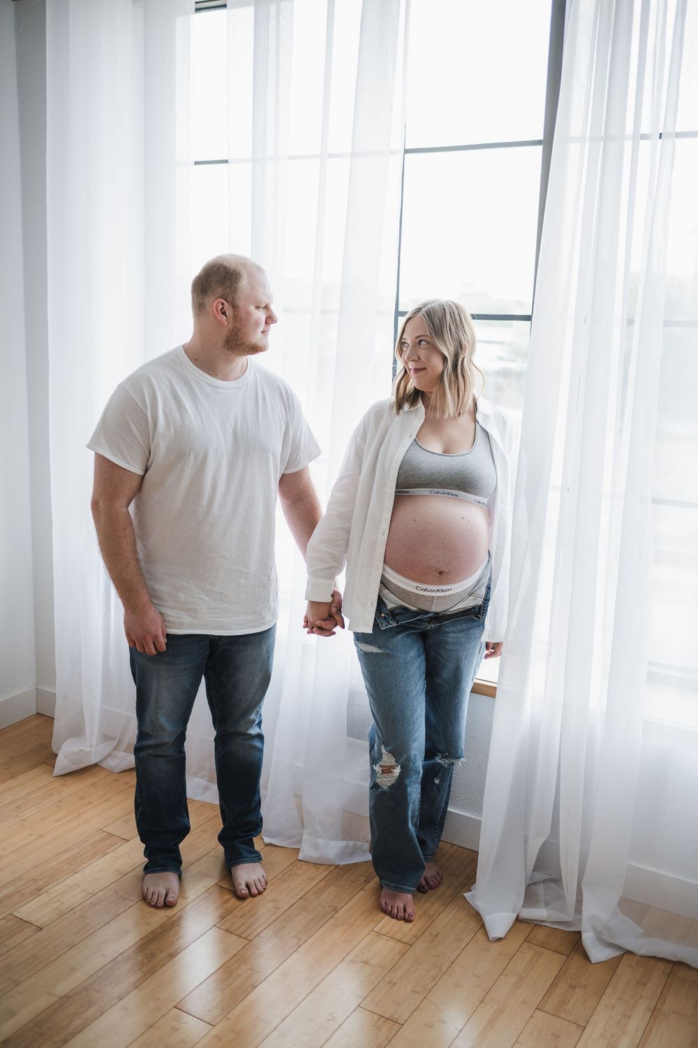 Expectant couple standing by sheer white curtains in a bright room.