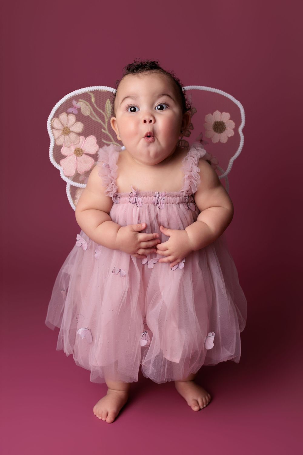 A baby in a pink tulle fairy dress with floral wings making a surprised expression against a burgundy background.