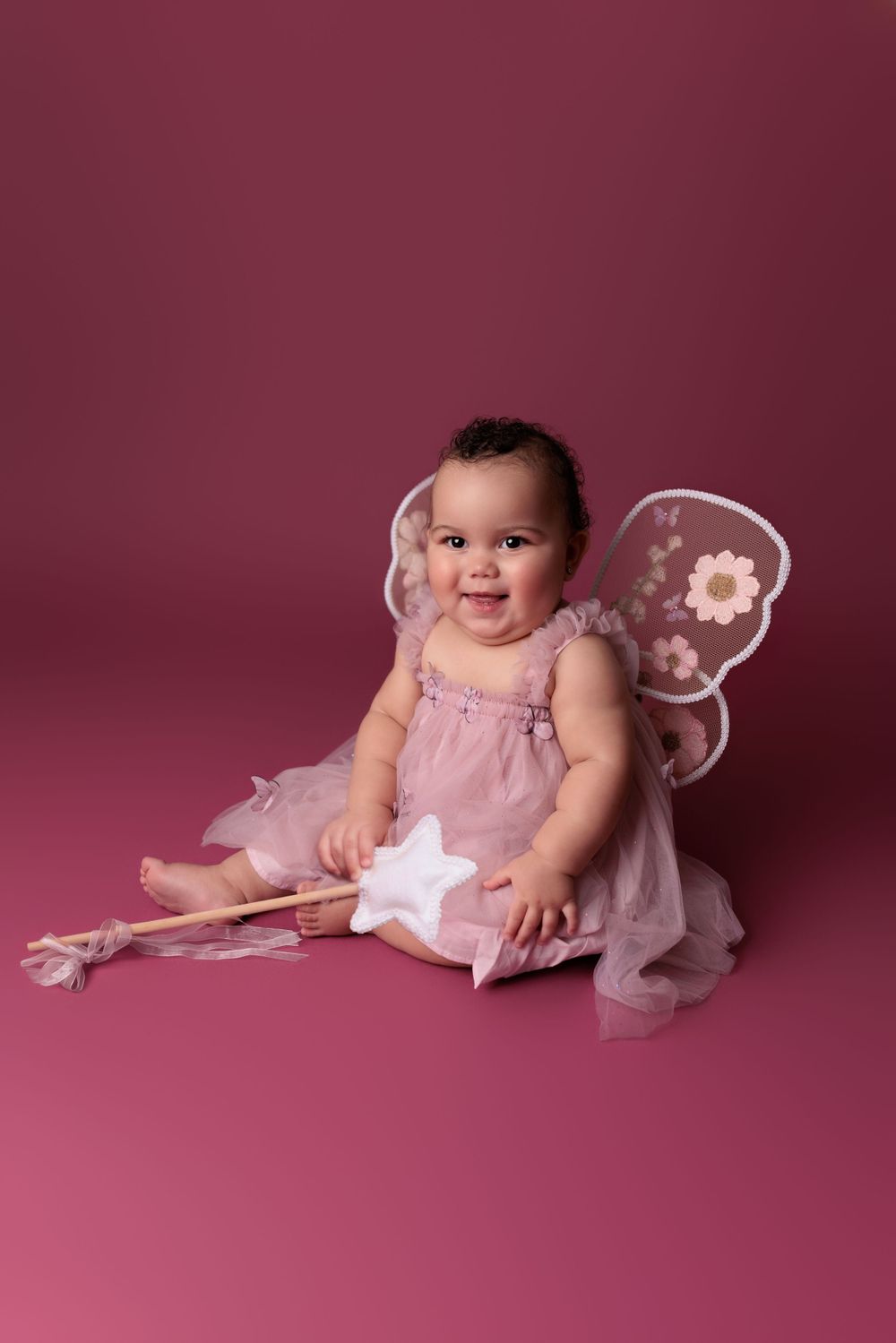 A baby wearing a pink tulle dress and fairy wings sits smiling on a pink backdrop, holding a decorative star wand.