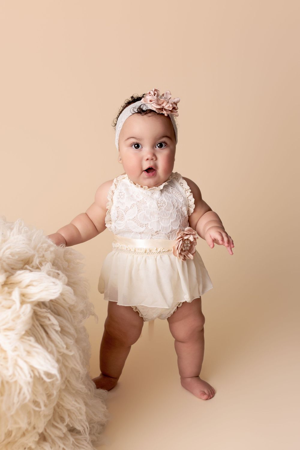 Baby wearing white lace romper with floral headband stands against beige backdrop next to cream-colored textured prop.