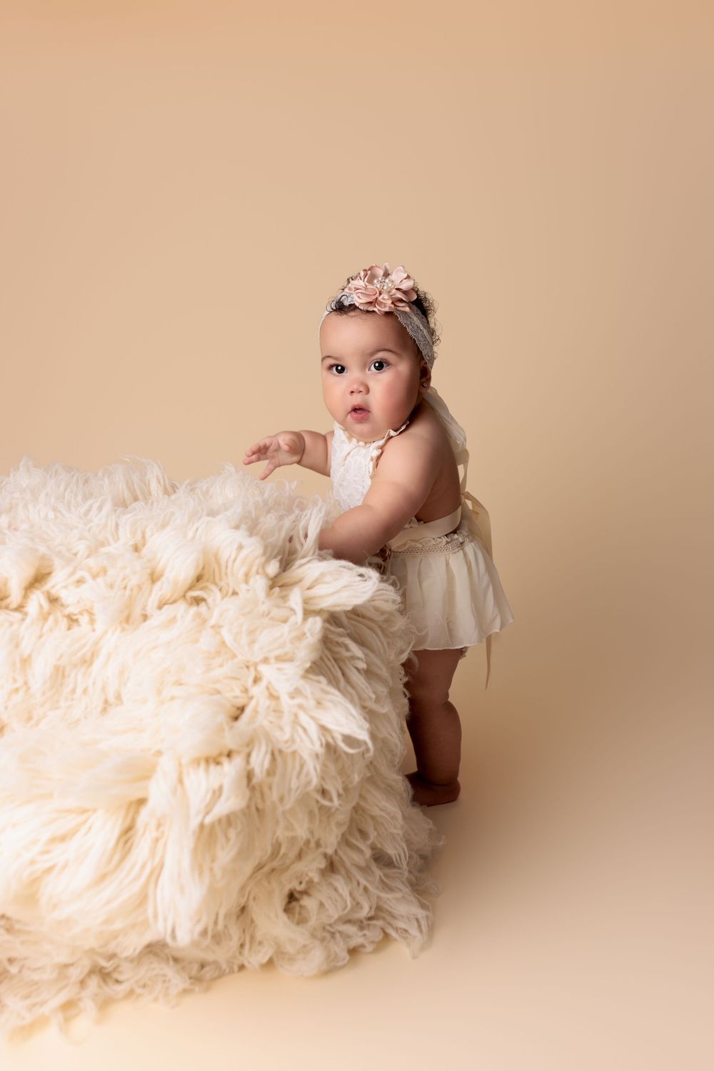 Baby in cream dress and floral headband stands beside fluffy white fur blanket against beige backdrop in studio portrait.