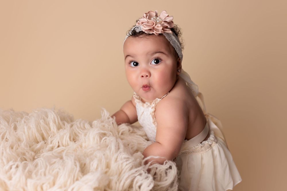 Sweet baby in white dress with floral headband poses on cream-colored textured blanket with surprised expression.