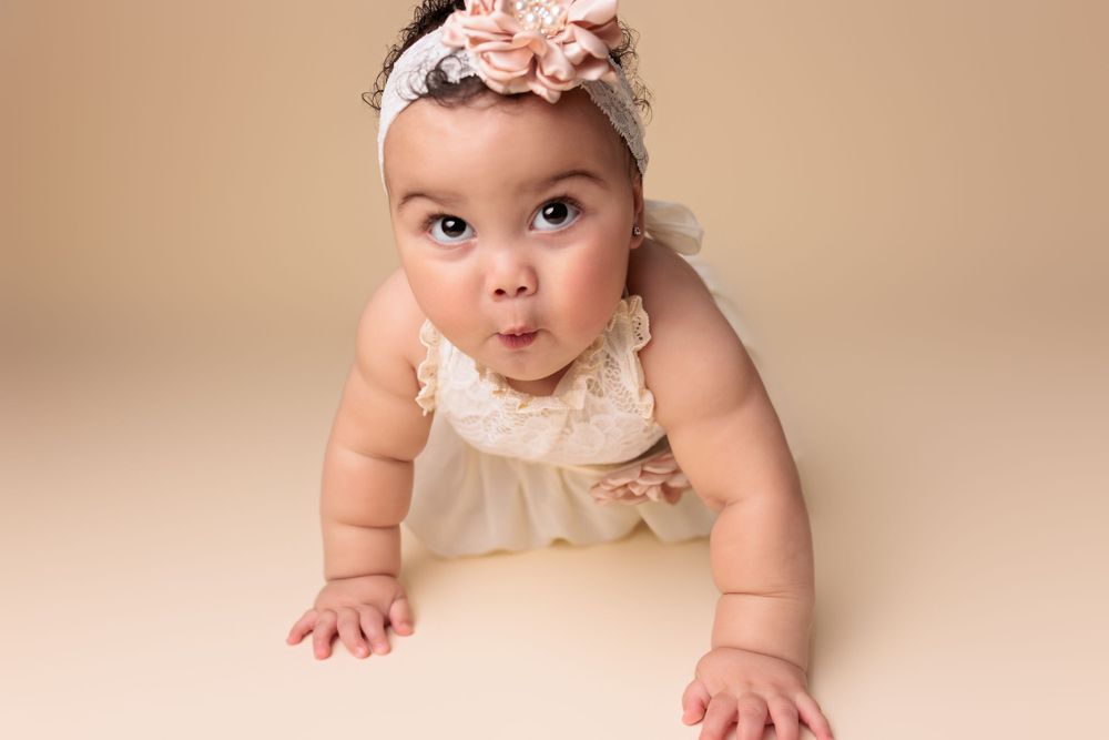 Baby in cream colored dress and floral headband making kissing face while crawling on beige backdrop.
