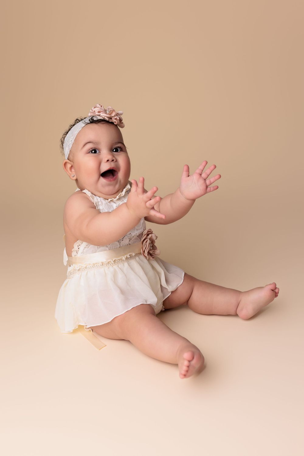 Baby wearing white dress and floral headband sits on beige backdrop while excitedly clapping and smiling.