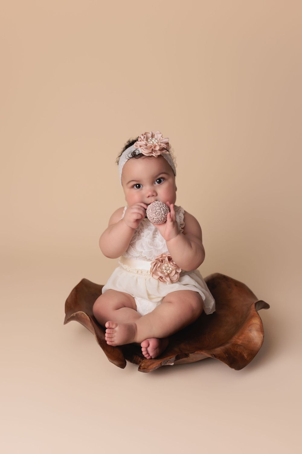 Baby in white lace romper with floral headband sits on brown leather while holding a small toy ornament on beige backdrop.