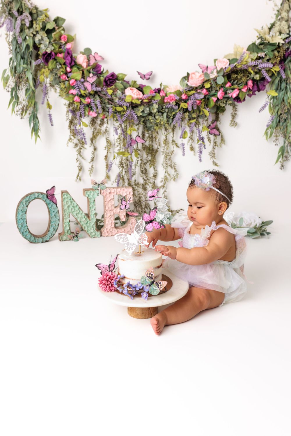 Baby in white dress sits with cake under floral garden arch with mint green 'ONE' letters for first birthday photoshoot.