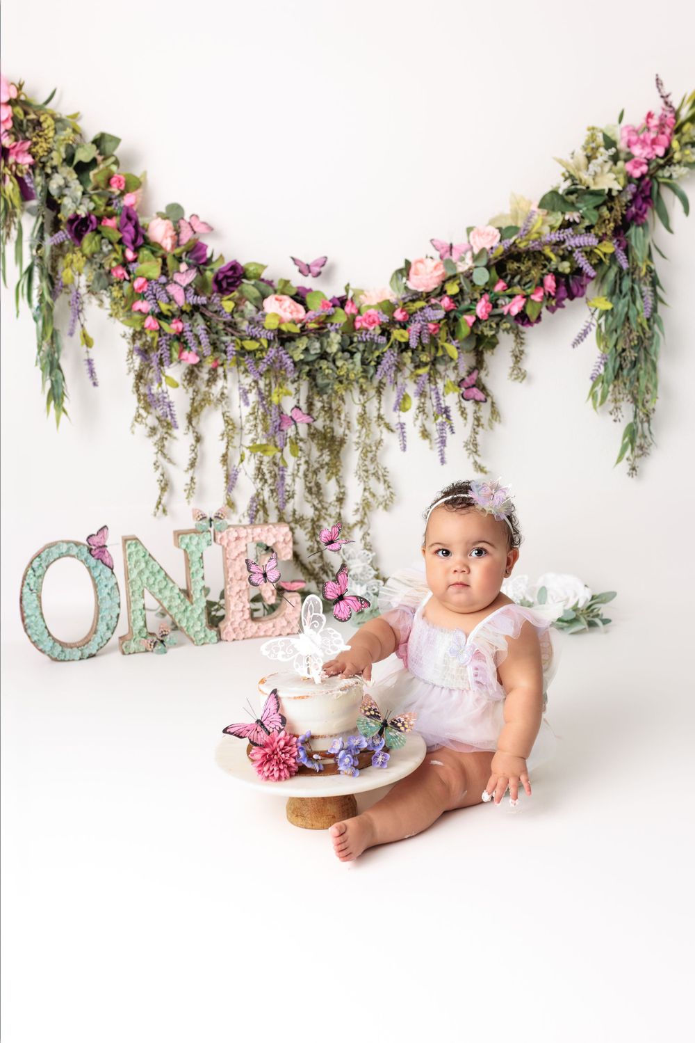 Beautiful floral garland with pink and purple flowers decorates a white backdrop with turquoise 'ONE' text for celebration setting.