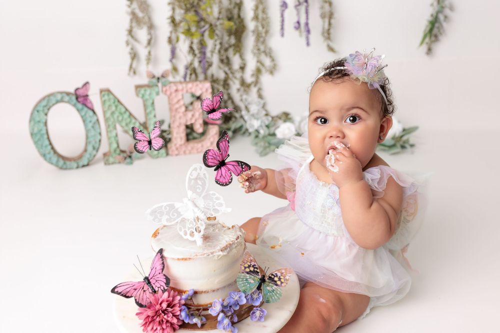 First birthday cake smash photo with pink butterflies and sparkly 'ONE' letters against white backdrop.