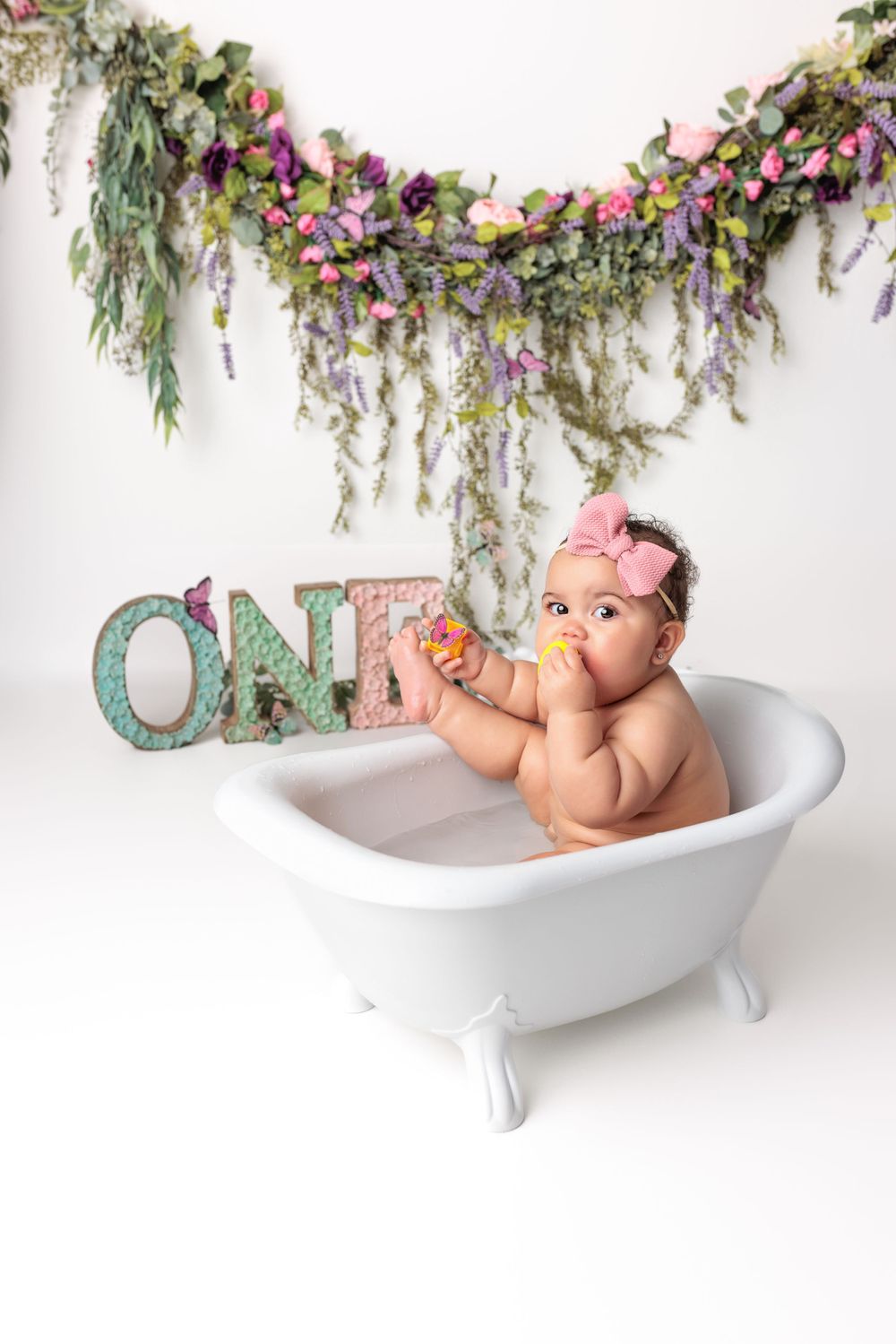 Miniature white bathtub decorated with floral garland and turquoise 'ONE' letters for baby milestone photography.