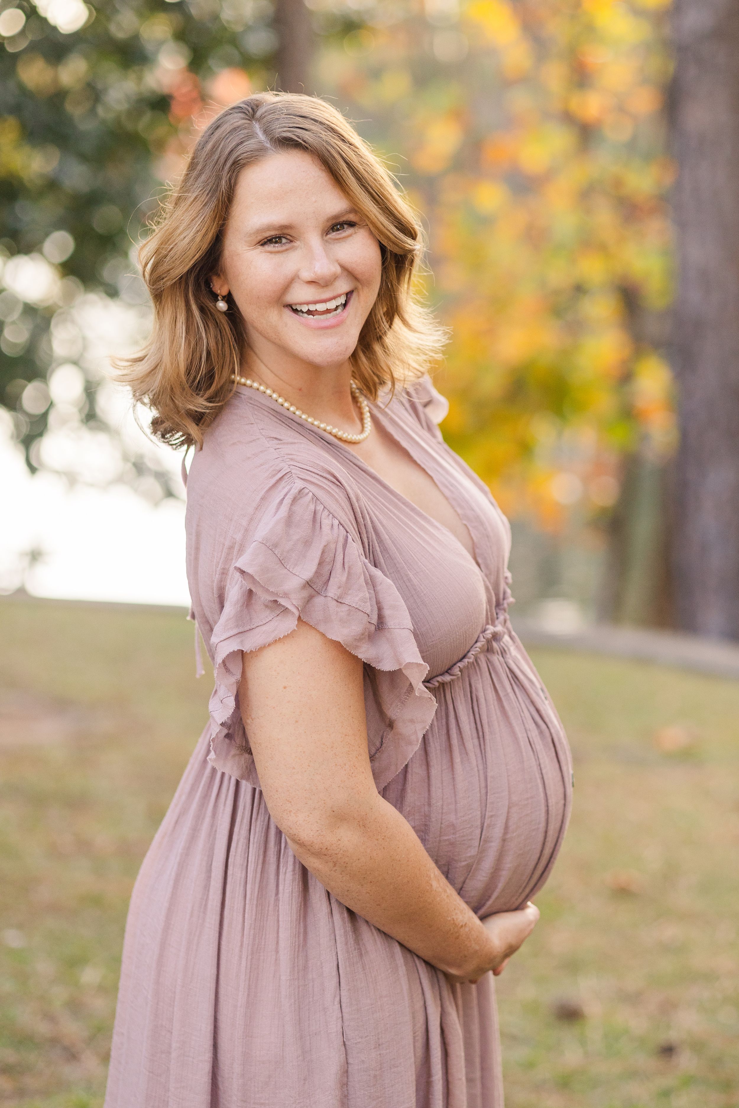 A person in a dusty rose ruffle sleeve dress poses outdoors against a backdrop of autumn foliage and bokeh lighting.