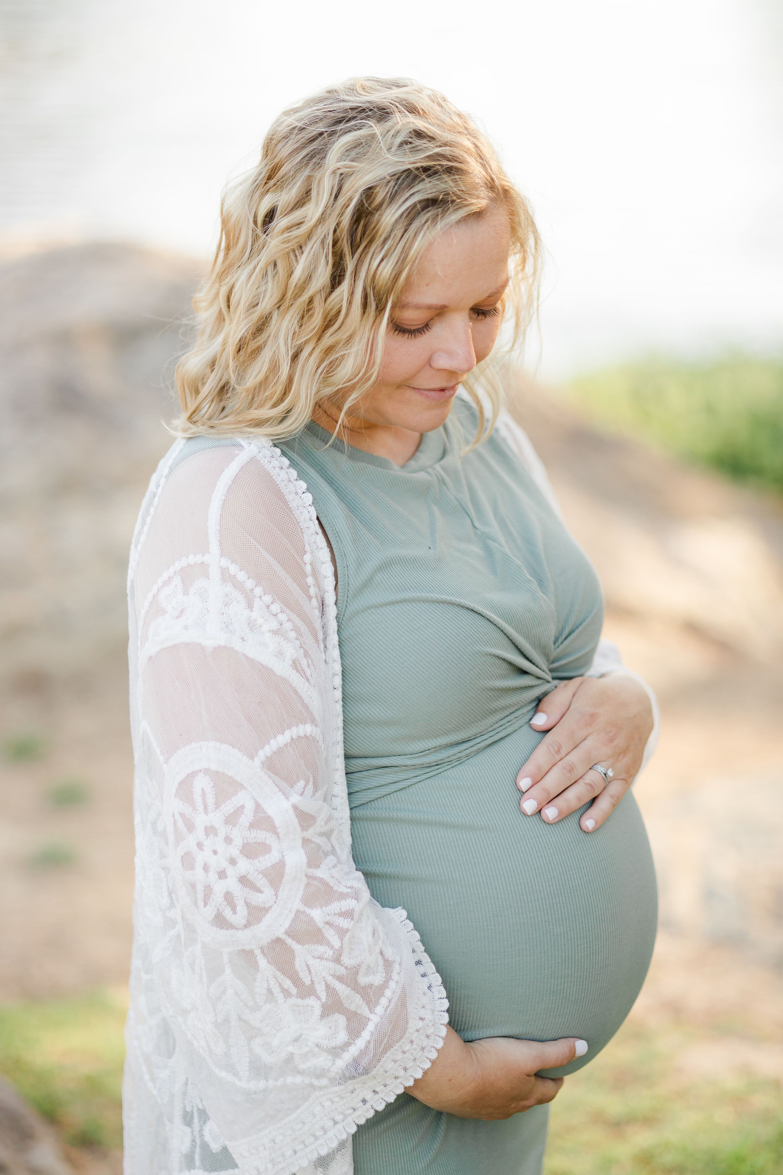 Pregnant woman in sage green dress and white lace shawl touches her belly during outdoor maternity photoshoot.
