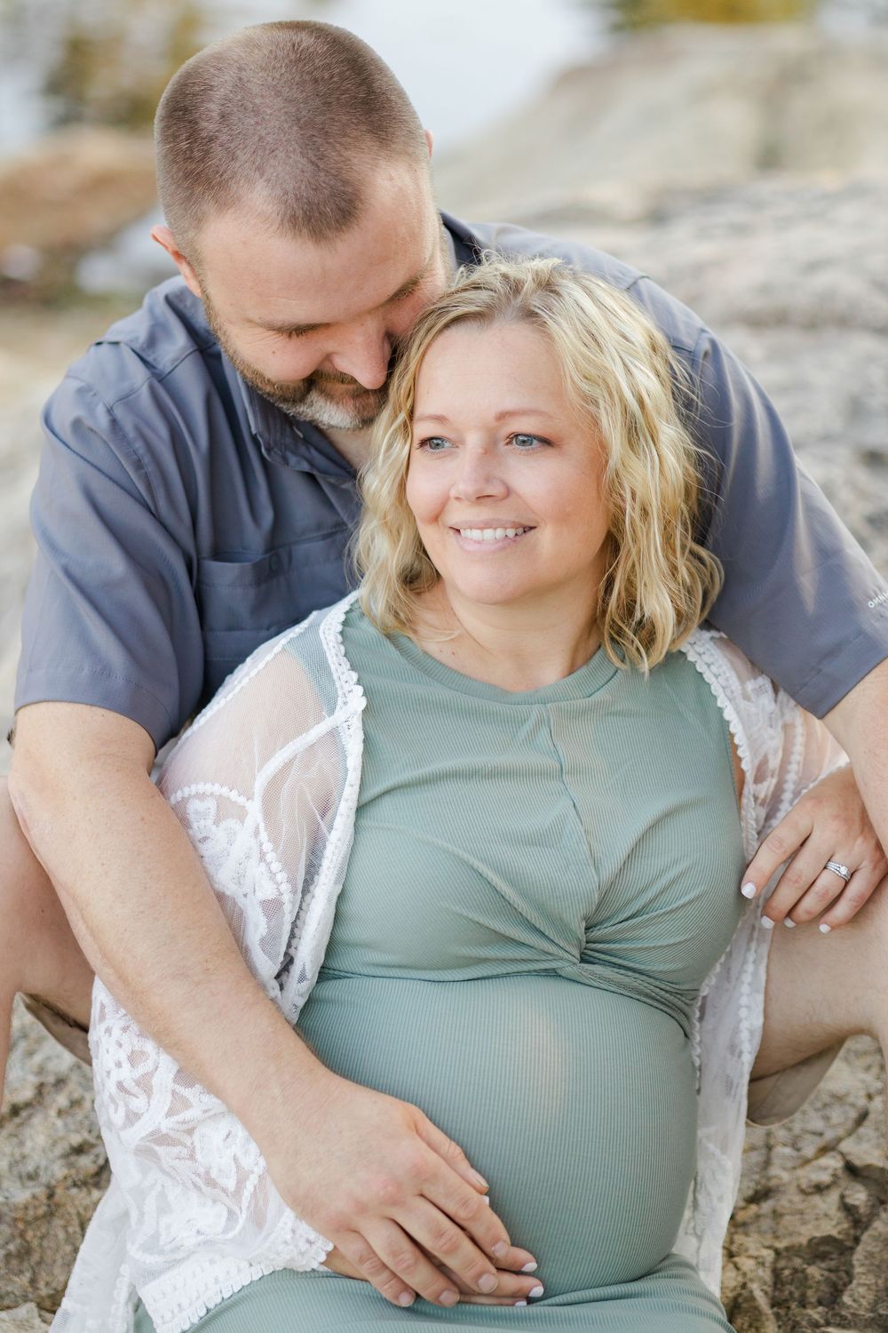 A couple shares tender moments together while resting on rocks during a maternity photoshoot outdoors.