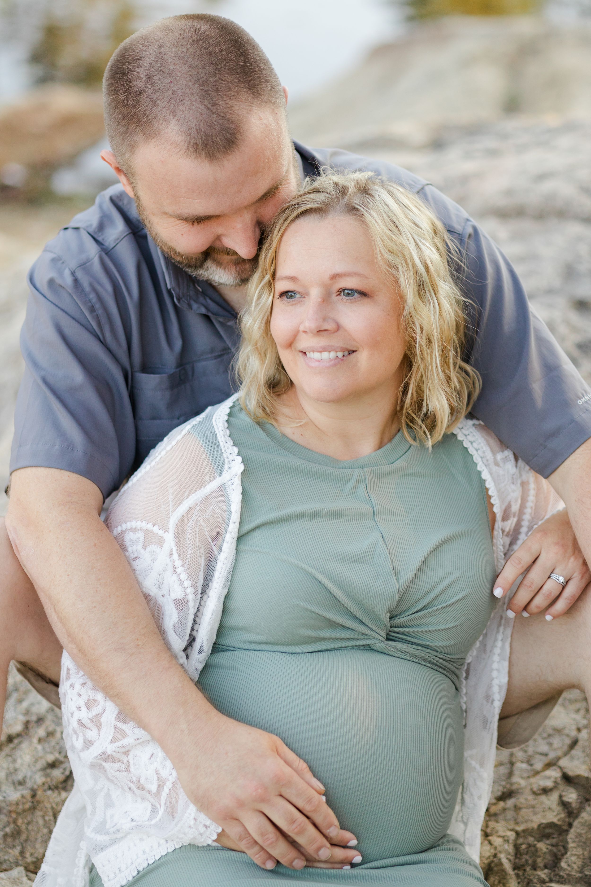 A couple shares tender moments together while resting on rocks during a maternity photoshoot outdoors.