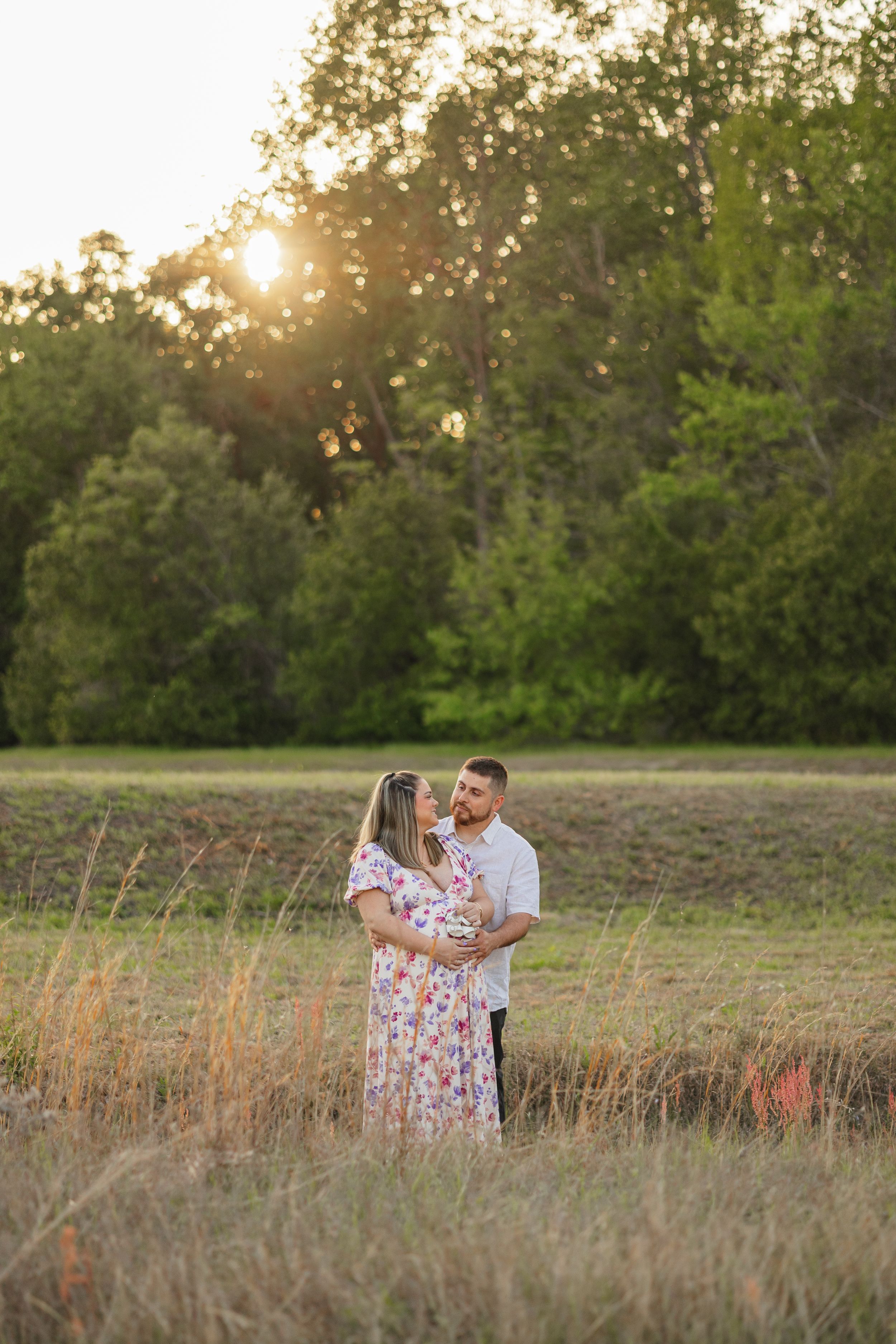 A couple stands together in a golden field at sunset with trees in the background.
