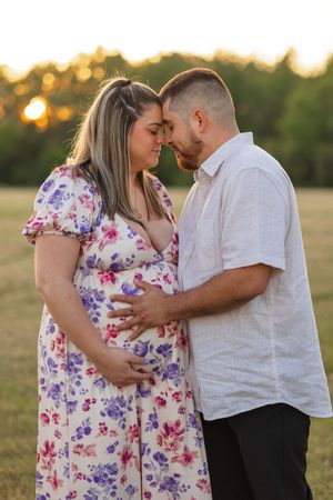 Romantic couple in floral dress and white shirt embrace during golden hour outdoor photoshoot.