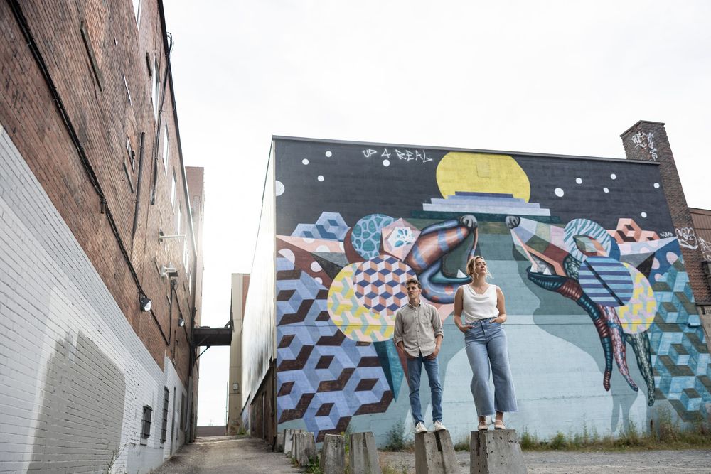 A engaged couple poses in front of a colorful geometric mural painted on a brick wall in an urban setting.  aKaiserPhoto.