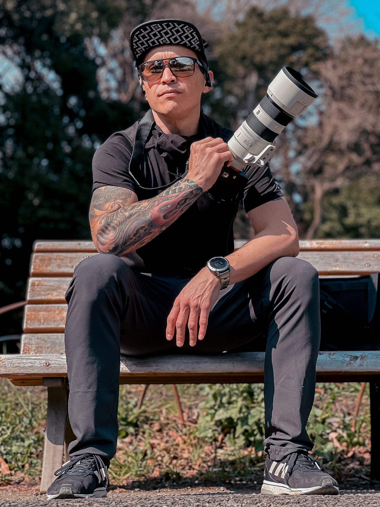 Professional photographer sitting on park bench with telephoto lens wearing casual attire and bandana headwear.